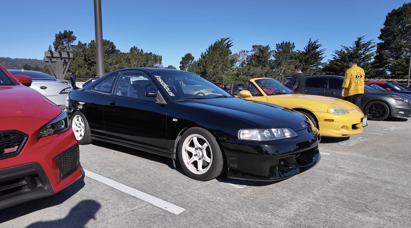 Modified black Honda Integra with white wheels beside a red Subaru STI at a Cars and Coffee meet in Monterey