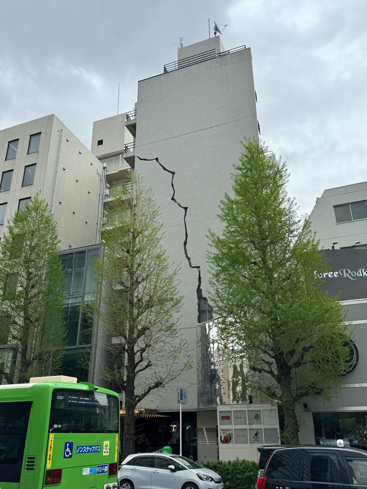 A tall concrete Harajuku building with a dramatic crack-line design, trees and a bus below